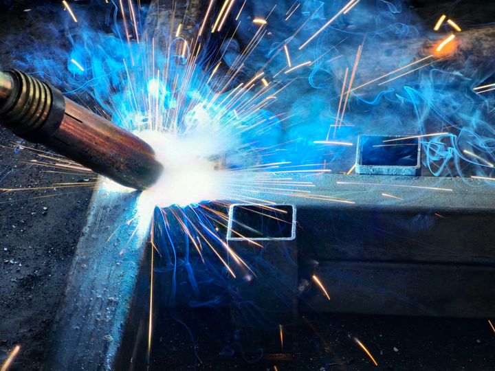 A close-up of a welding torch melting metal, creating bright blue light and flying sparks in a workshop.
