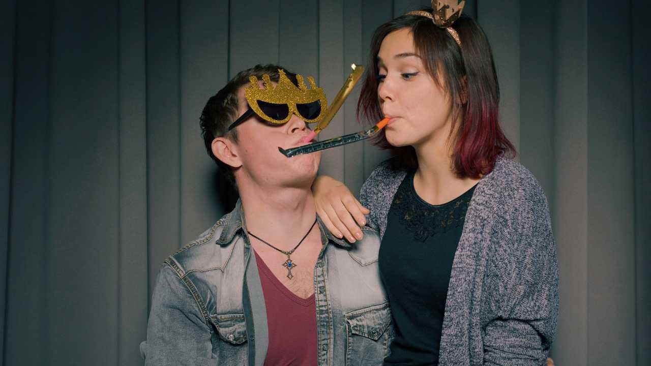 A man and a woman are blowing party horns in a photo booth.