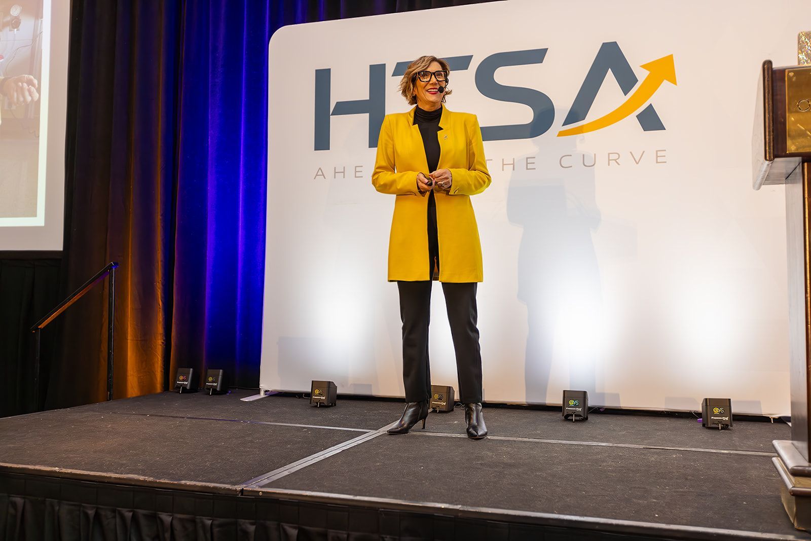 A woman in a yellow jacket is standing on a stage in front of a htsa logo