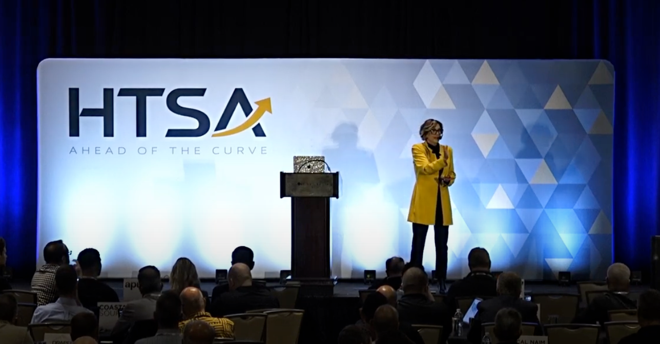 A woman in a yellow coat is giving a speech at a htsa conference.