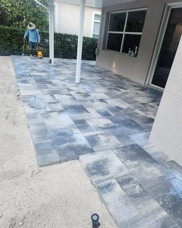 A person standing on a newly installed gray-toned paver patio next to a house under a covered porch.