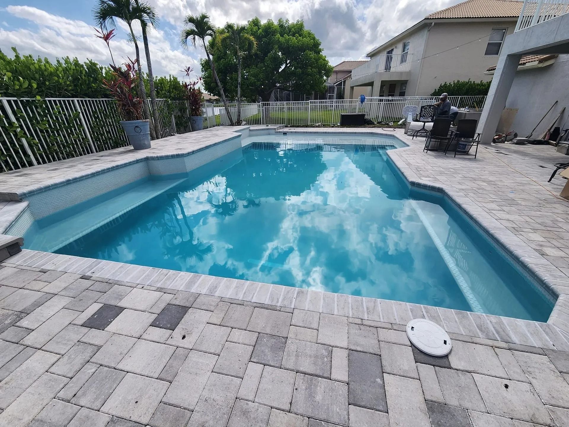 A rectangular swimming pool with blue water and light-colored paver decking, set in a backyard near a residential house.