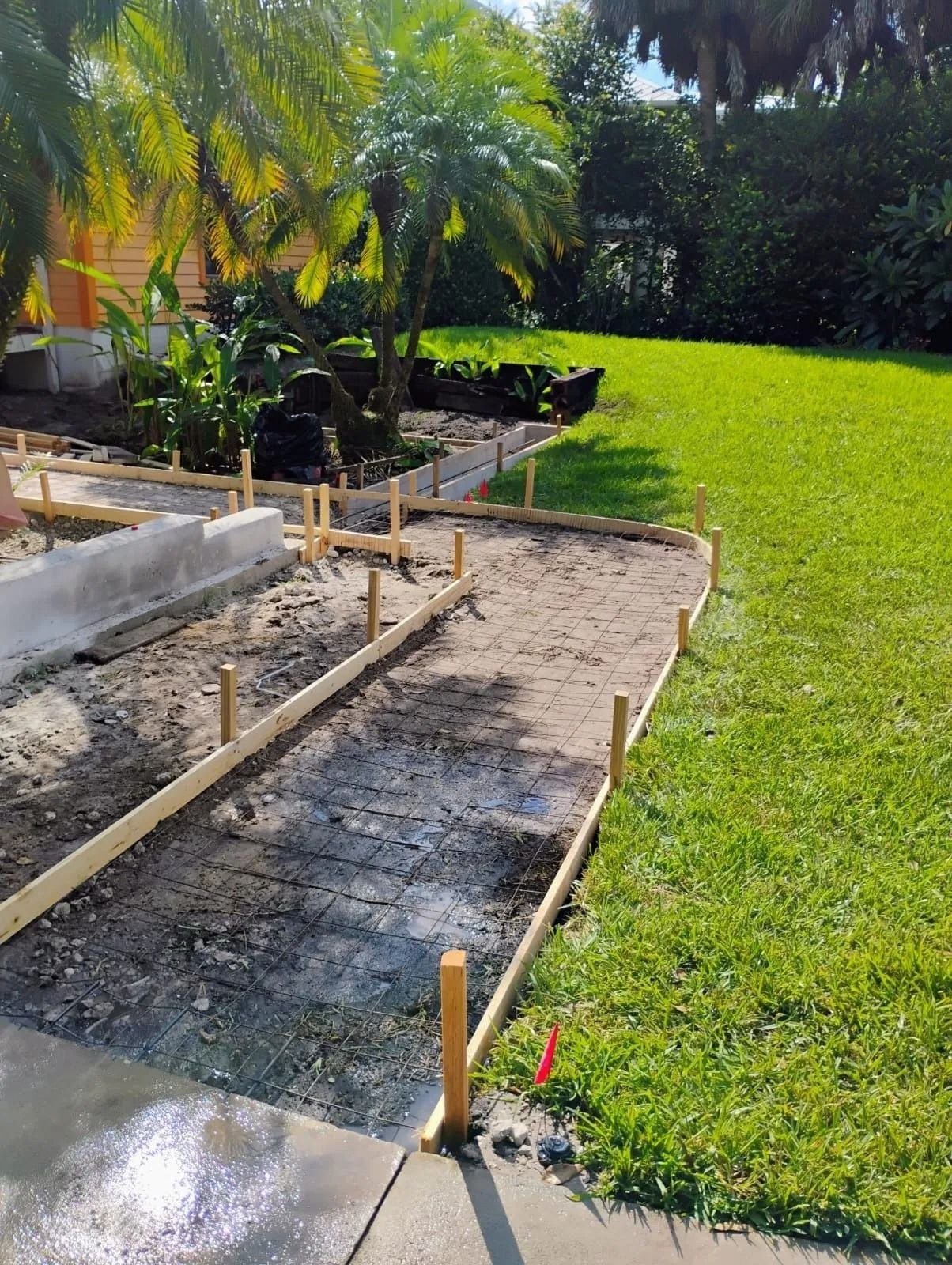 Wooden forms staked into the ground prepare a curved walkway for concrete pouring next to a grassy lawn.