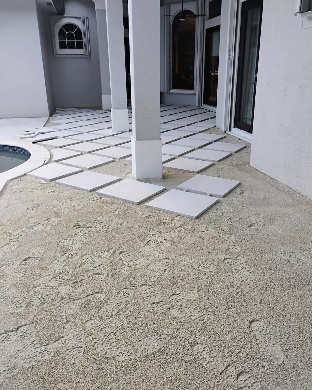 A modern patio featuring a textured sandy surface with light-colored square paver stones laid in a grid pattern.