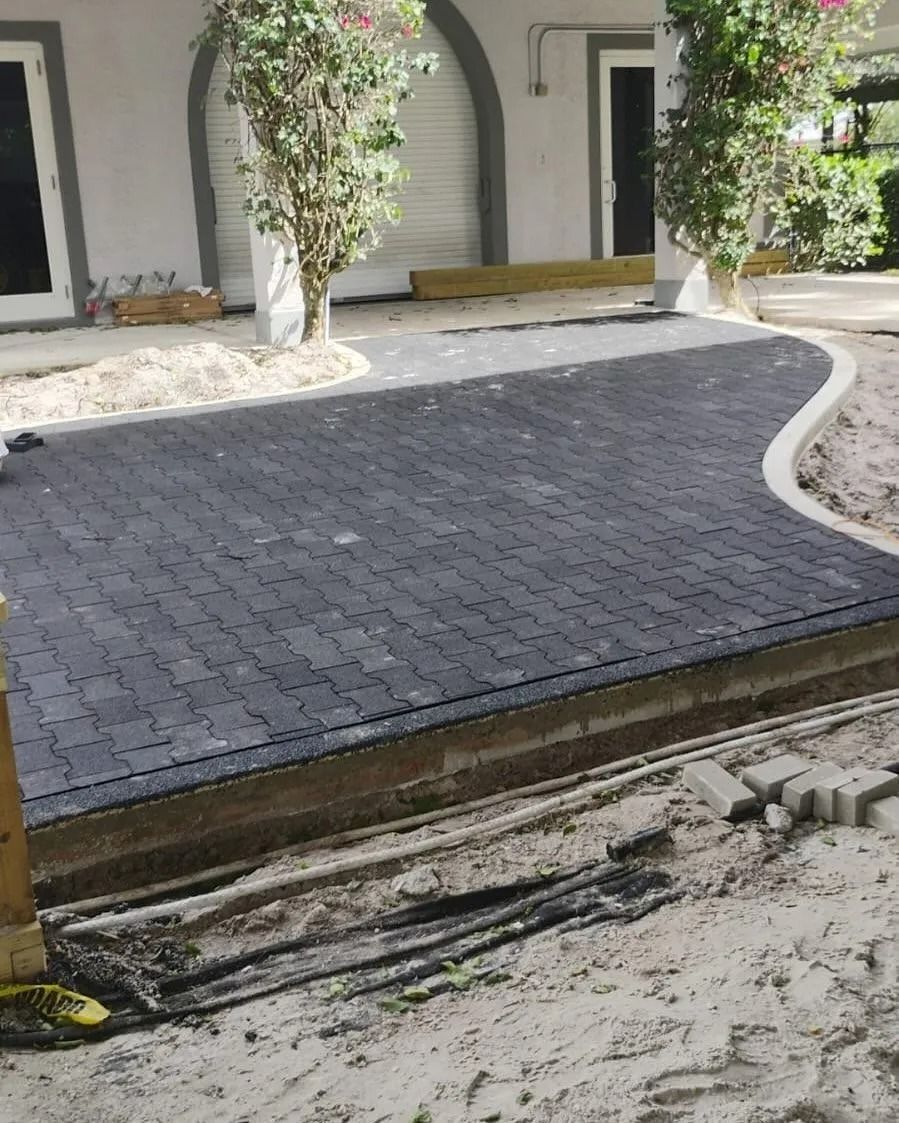 A curved, charcoal-colored brick paver patio sits unfinished in a sandy yard next to a house with two trees.