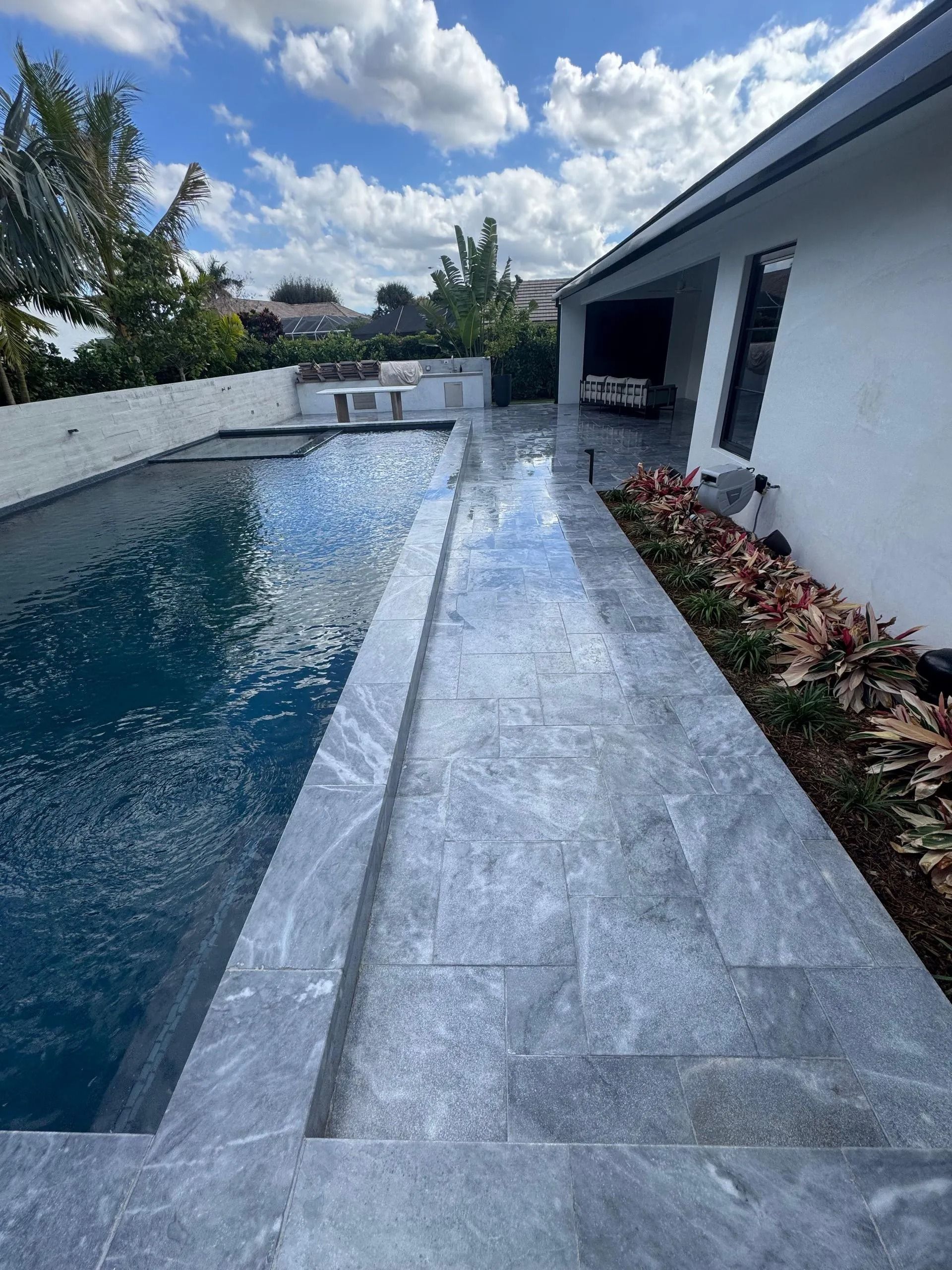 A swimming pool with grey stone pavers, a white house wall, and a flower garden under a blue sky with white clouds.