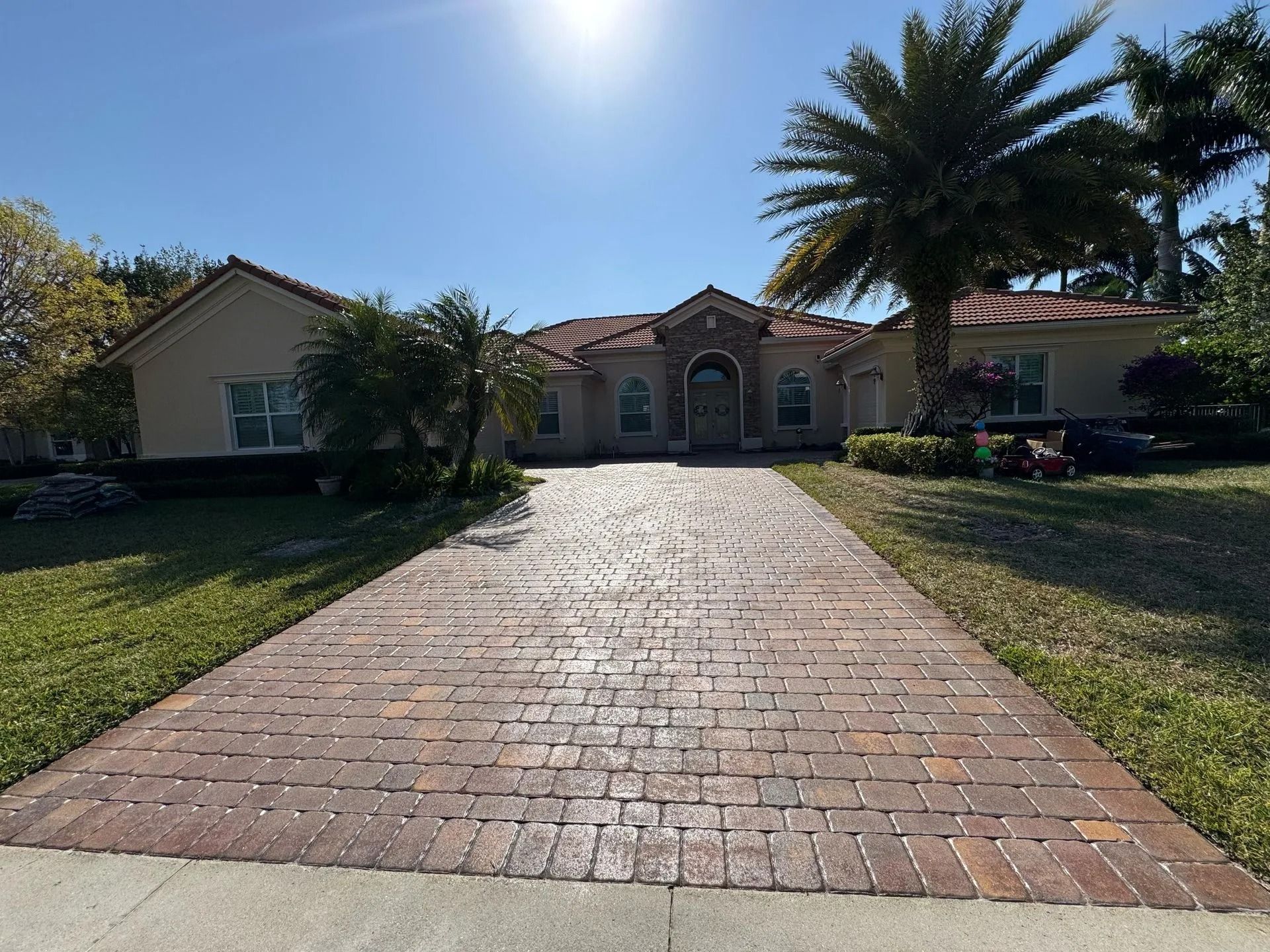 A sunlit single-story stucco house with a red tile roof and a long, patterned brick paver driveway.