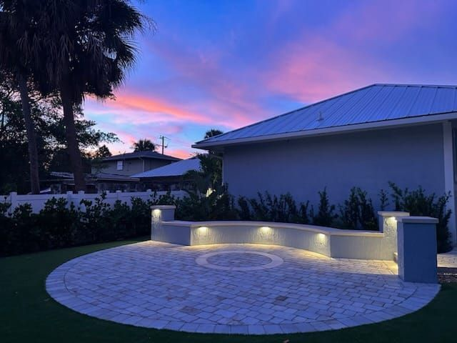 A lit, curved stone bench on a circular paved patio at sunset with purple and orange skies in the background.