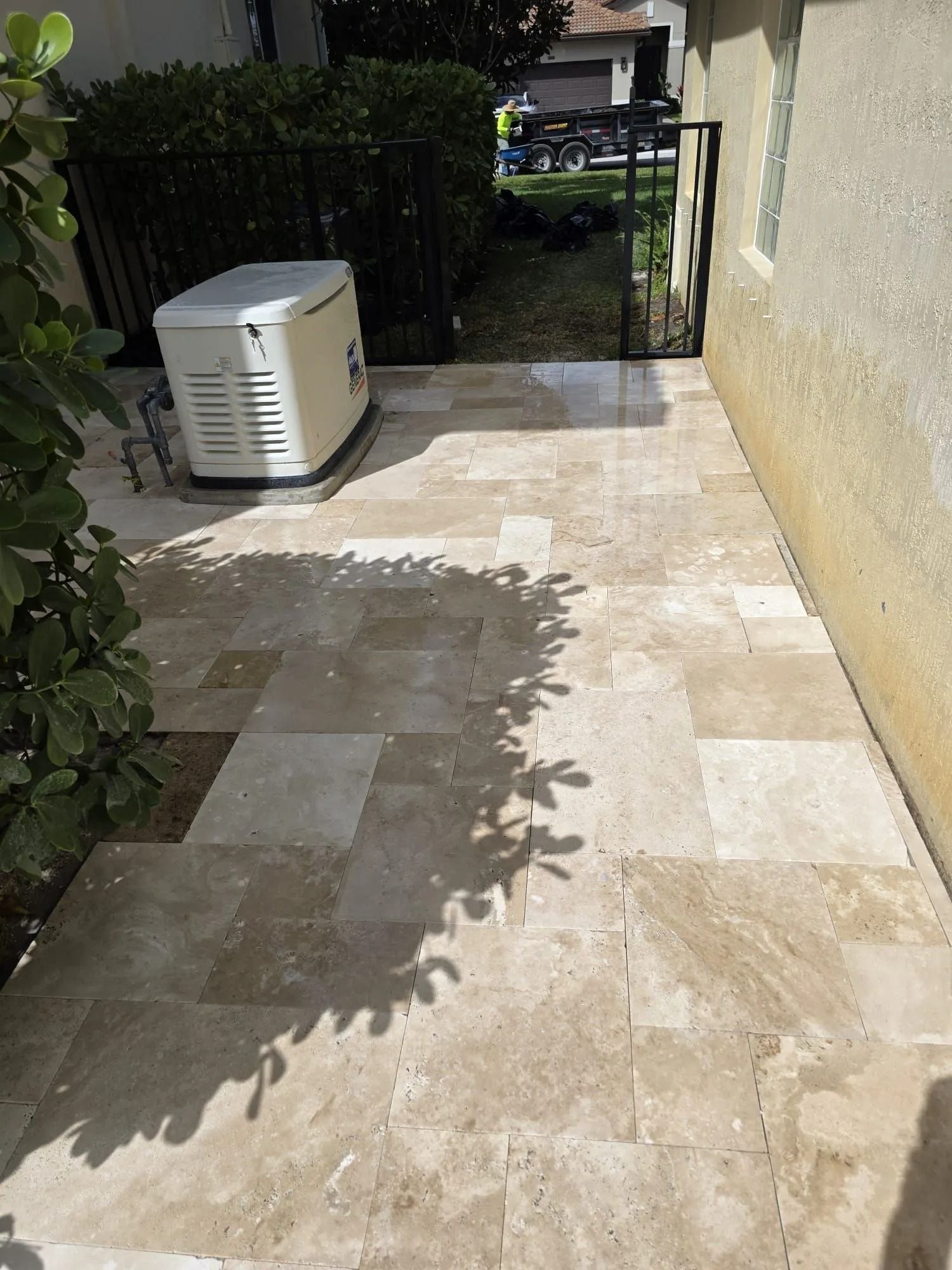 A paved patio made of light tan stone tiles, with a white generator sitting near a black metal fence and house wall.