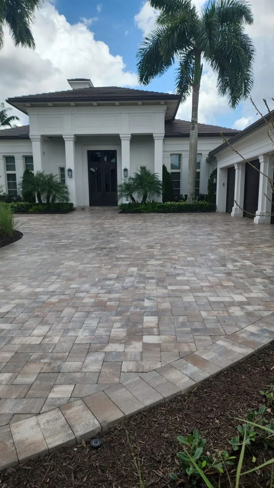 A bright, one-story white house with a dark front door, stone paver driveway, and a tall palm tree under a blue sky.