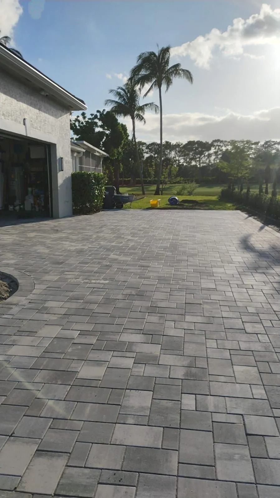 A gray paver driveway leads to a garage, with palm trees and a green yard in the background under a blue sky.