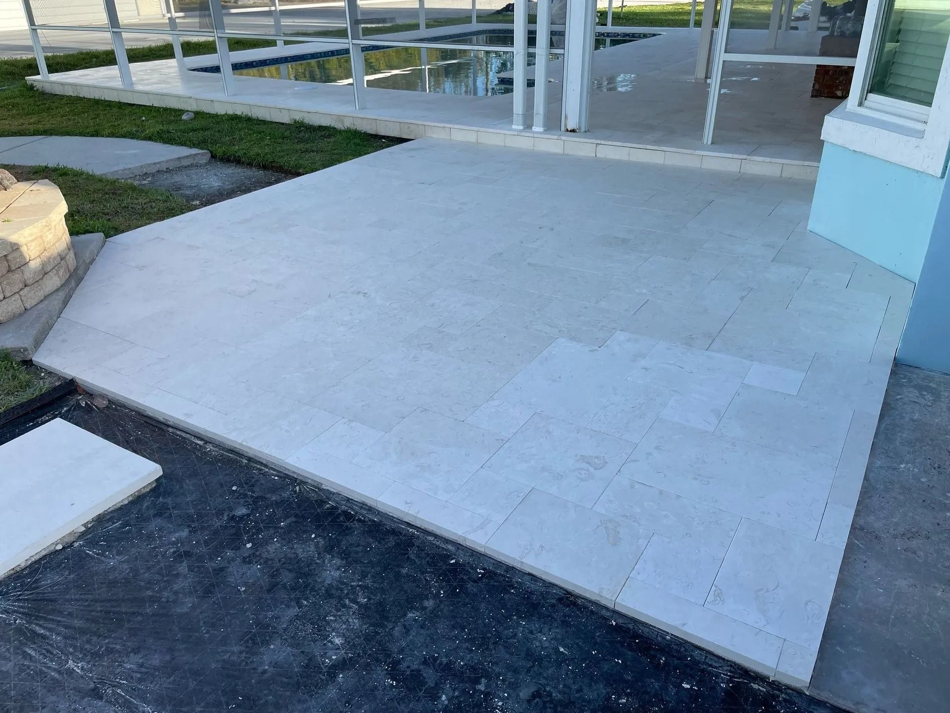 Newly laid light-colored stone pavers form a patio extending from a house toward a screened-in swimming pool area.