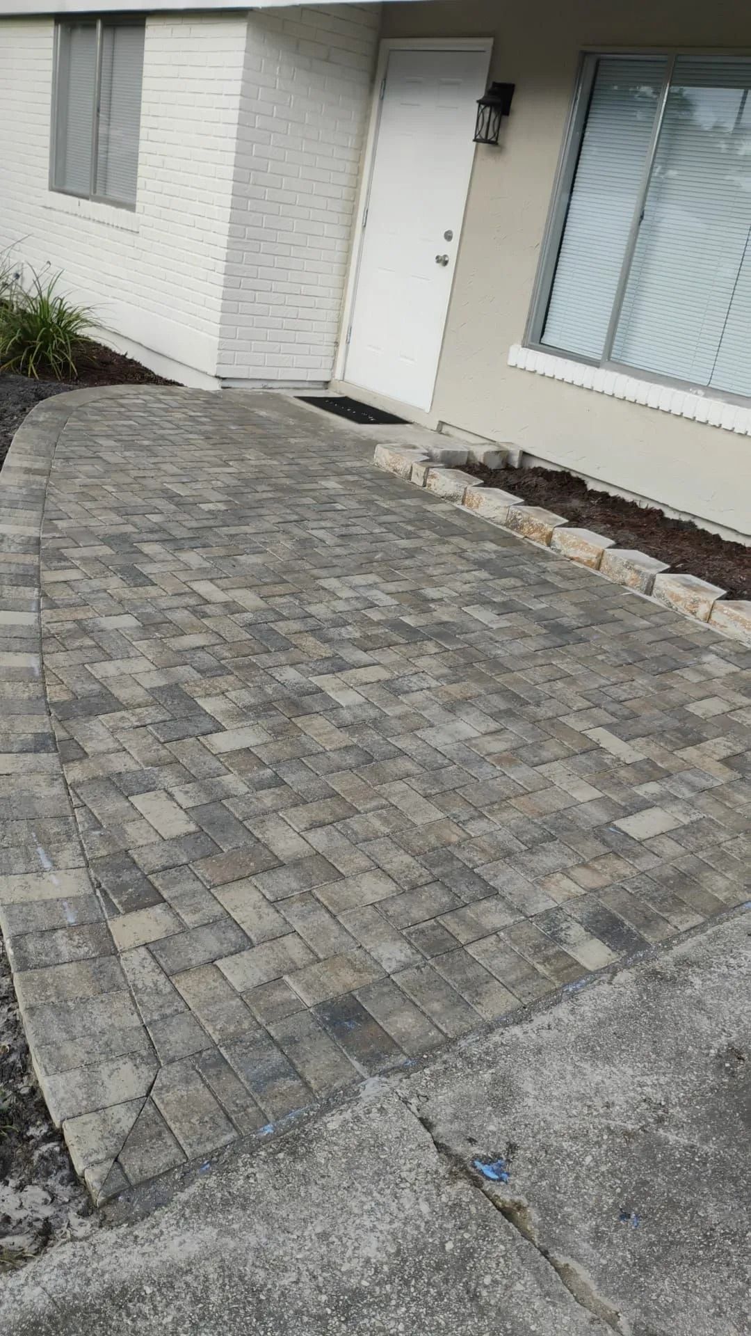 A newly installed gray paver patio leading to a white residential front door, bordered by stone edging.