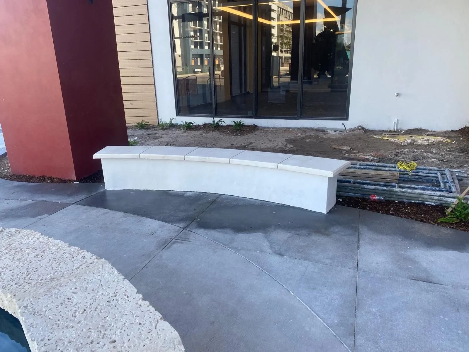 A curved, white concrete bench sits on a gray patio in front of a building with large glass doors.