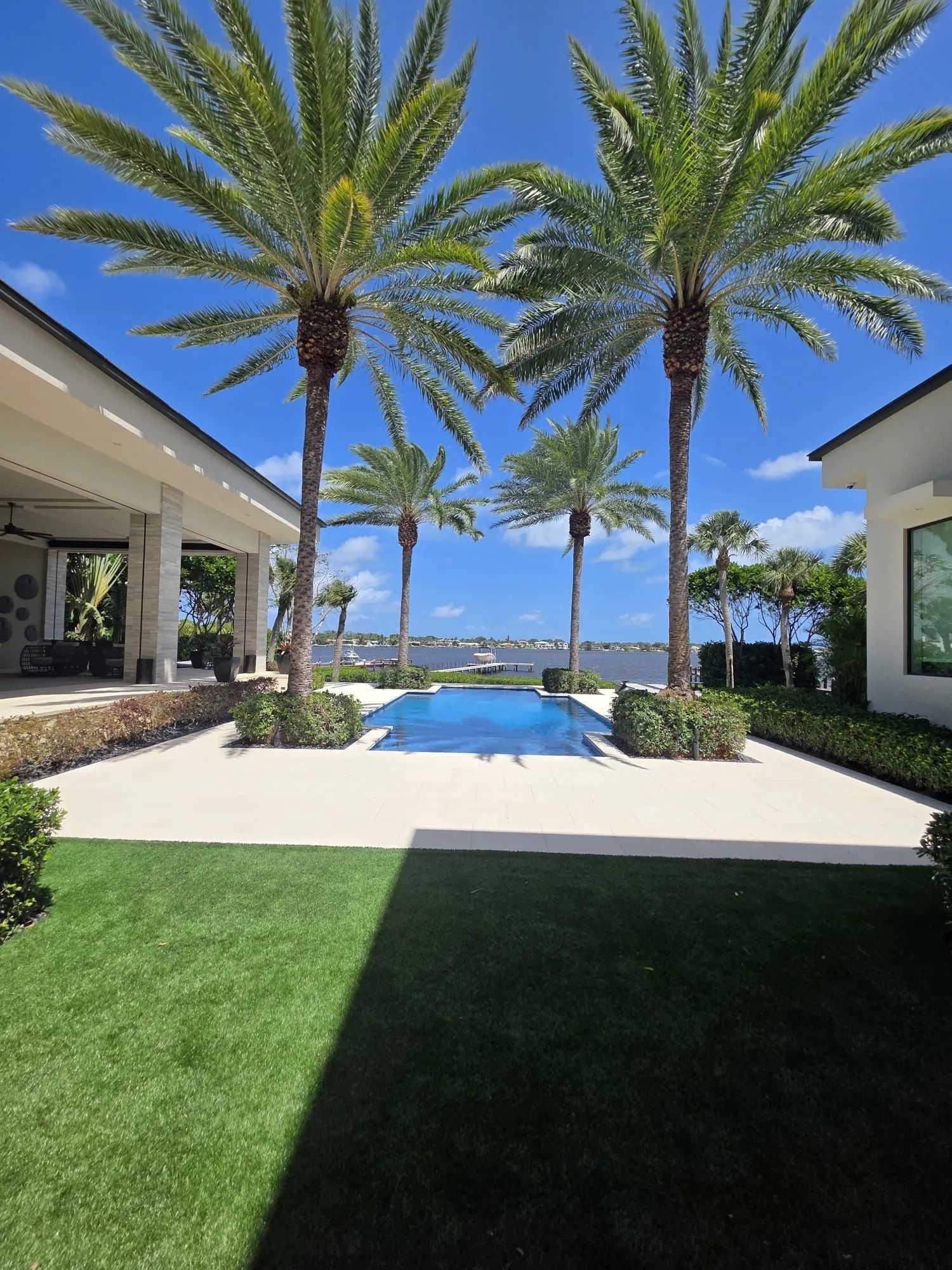 A luxury patio with a swimming pool looking out towards a waterfront vista, flanked by tall palm trees under blue skies.