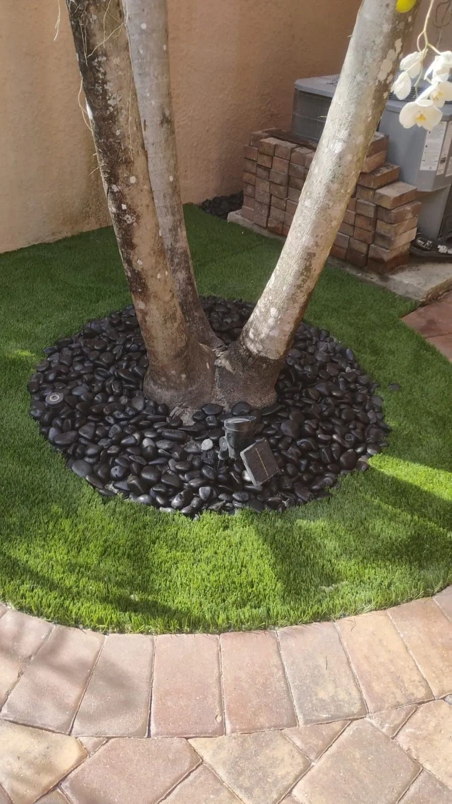 A tree trunk in a yard with a circular bed of black river rocks surrounded by artificial grass and paver stones.