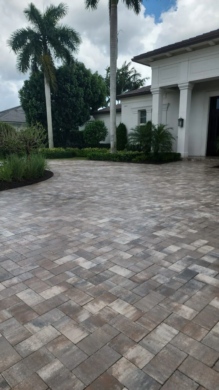 A paved driveway leads to a white house with columns, surrounded by palm trees and manicured green hedges.