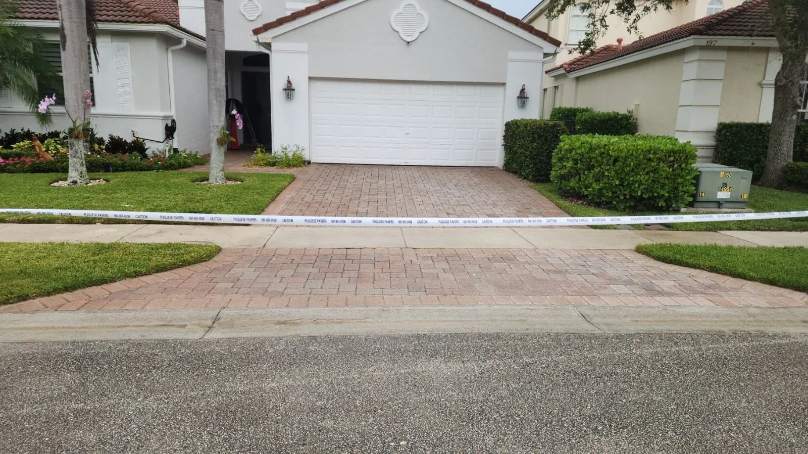 A suburban house with a brick driveway blocked by a strip of white police tape stretched across the sidewalk.