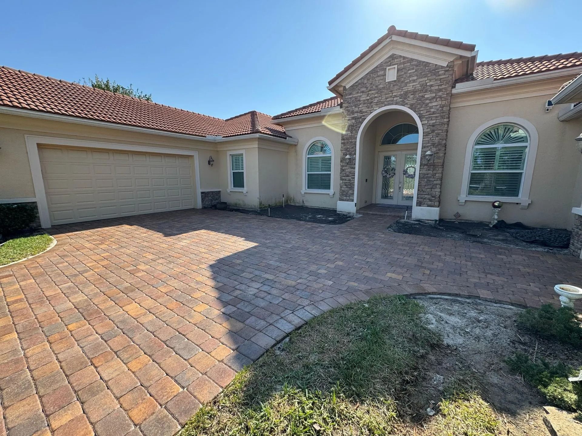 A single-story beige stucco house with a stone entryway, arched windows, a tile roof, and a paved brick driveway.