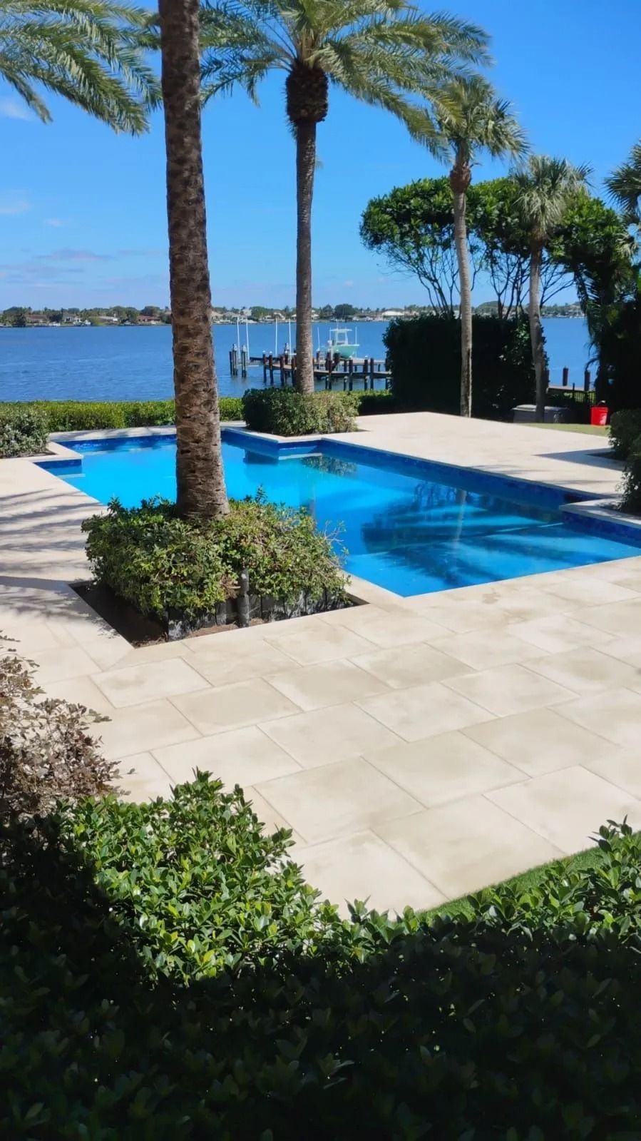A swimming pool surrounded by a light-colored stone patio, palm trees, and lush landscaping, overlooking a calm blue bay.