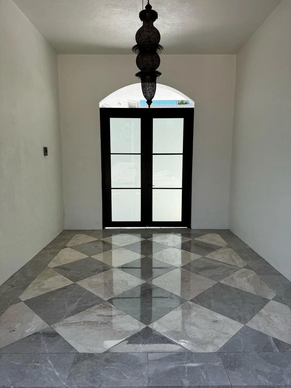 A foyer with white walls, a diamond-patterned gray and white marble floor, and an arched glass door beneath a pendant light.