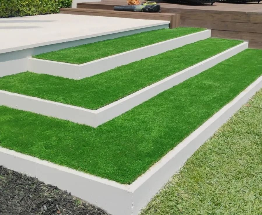 Three tiered garden steps with artificial grass, white borders, and a dark stone and natural grass surround.