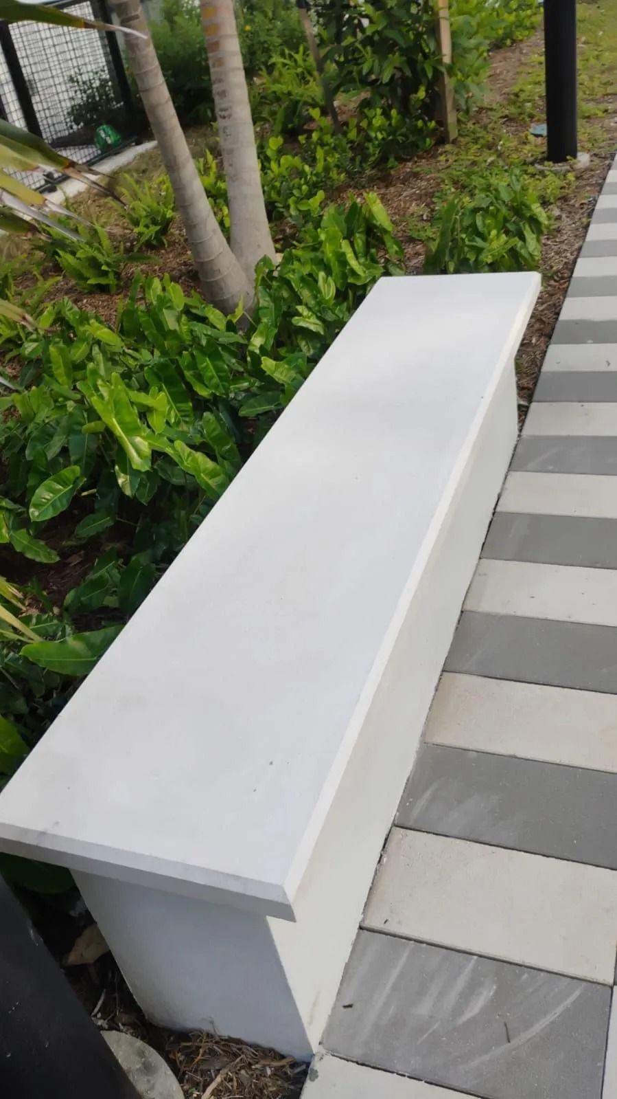 A long, white, rectangular concrete bench sits next to a path made of alternating gray and white pavers in a garden.