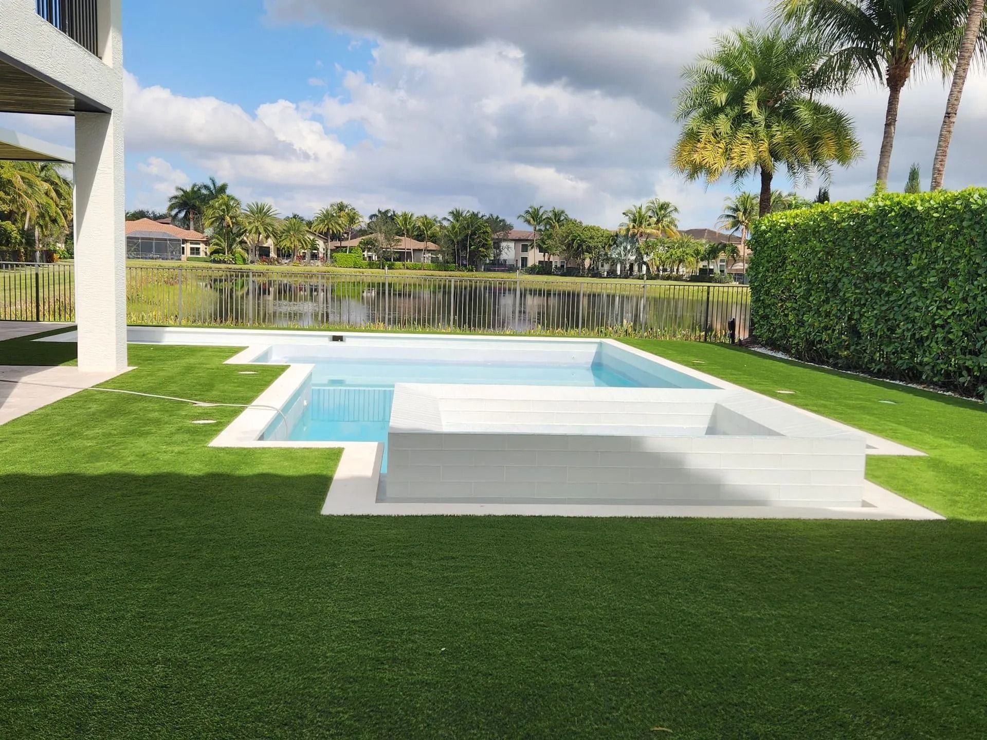 A modern pool with a shallow sun shelf and built-in steps, surrounded by green turf and a hedge, overlooking a lake.