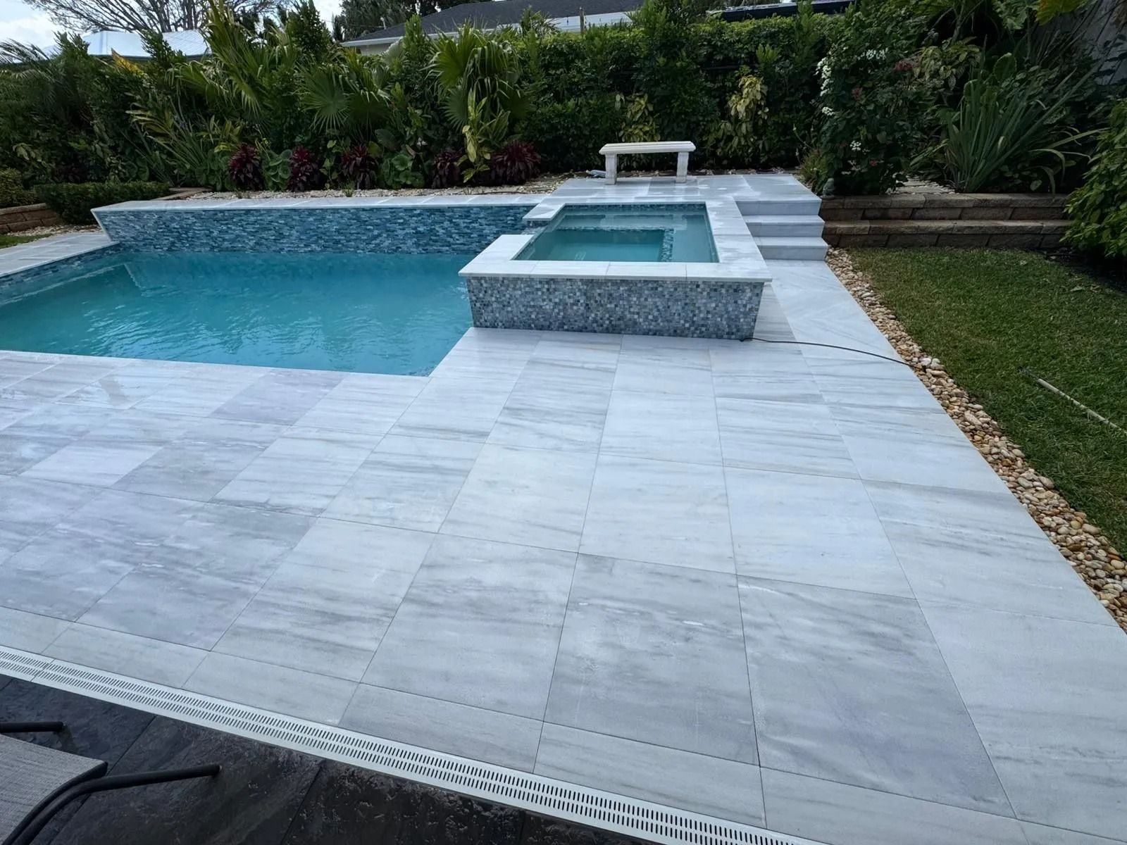 A rectangular swimming pool with an attached square spa, surrounded by light gray stone pavers and lush green landscaping.