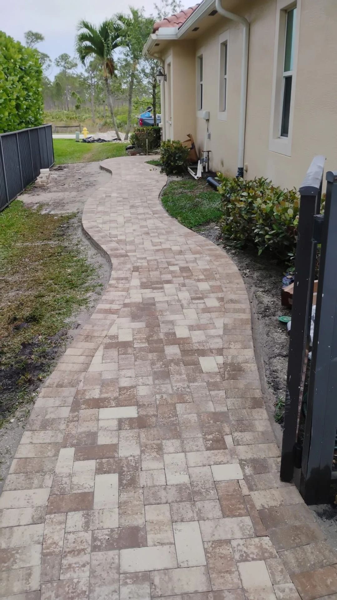 A winding walkway made of light and dark brown pavers leading alongside the side of a tan stucco house.
