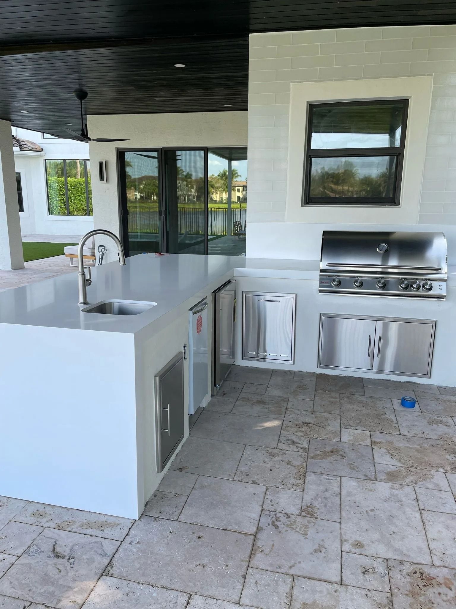 An outdoor kitchen with white counters, stainless steel grill, and sink on a stone patio under a black ceiling.