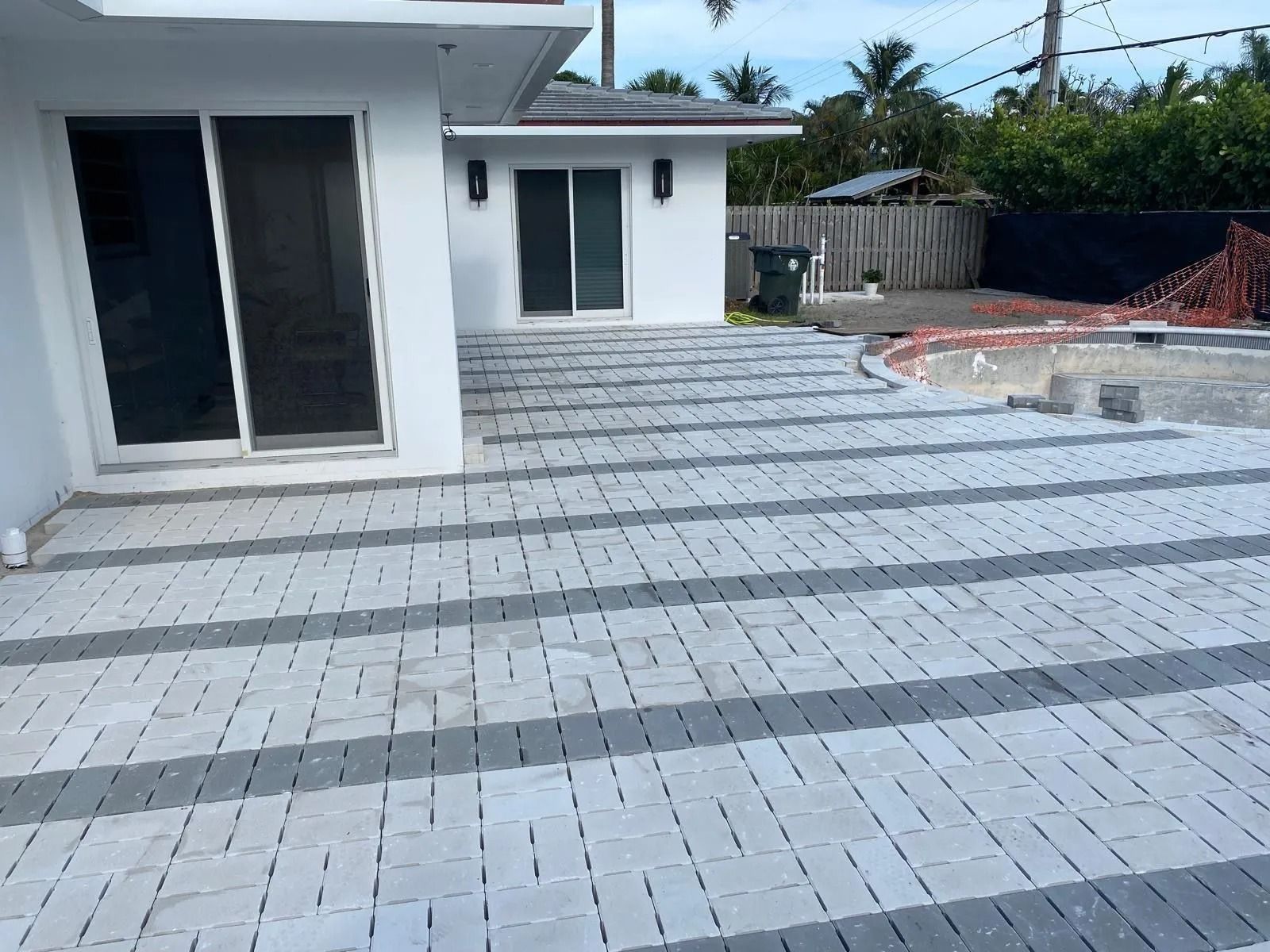 White house with sliding glass doors and a grey-and-white patterned paver patio leading to an empty pool area.