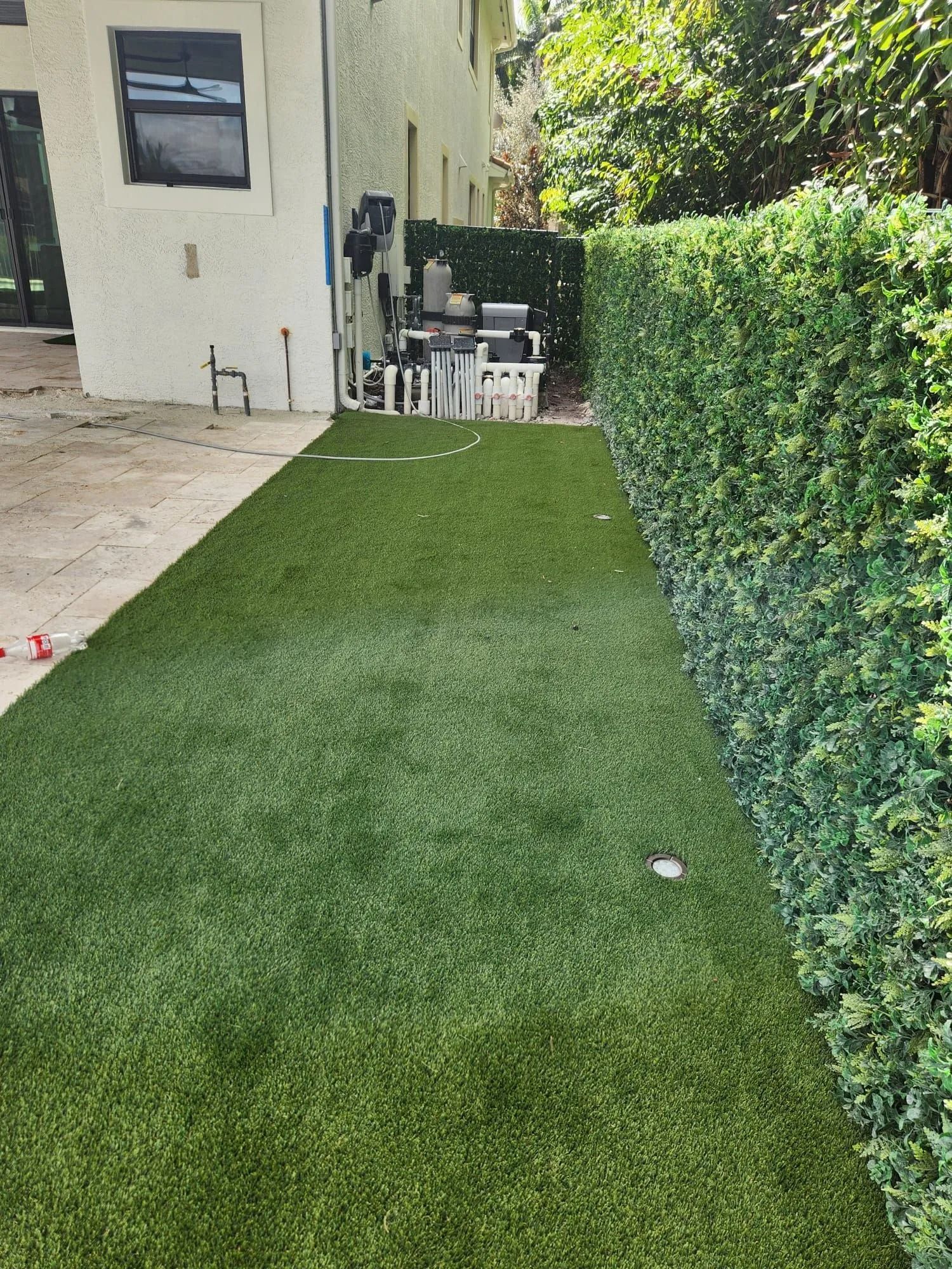 A narrow strip of artificial green grass leading from a house patio toward a tall green hedge.