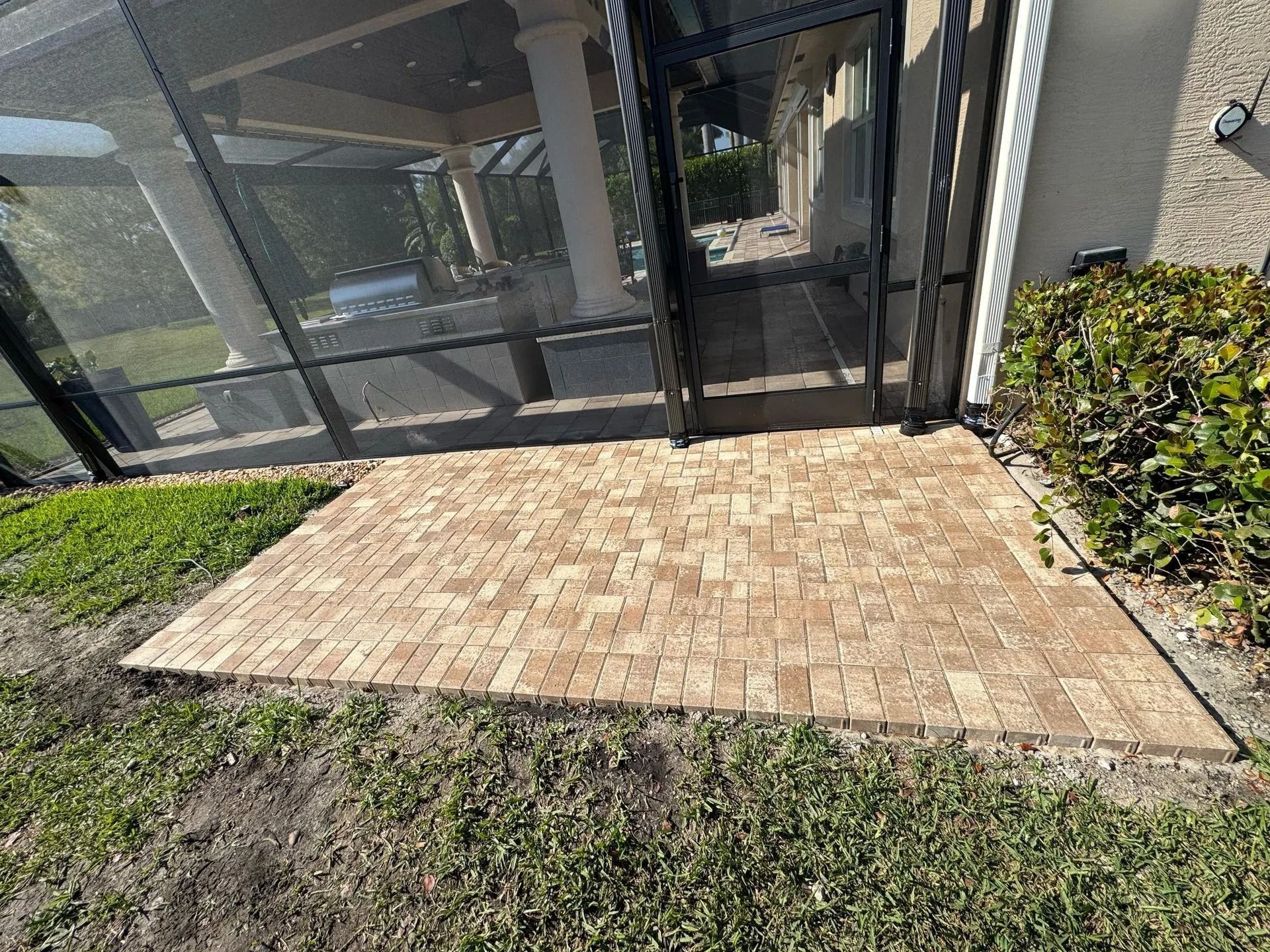A freshly installed rectangular brick paver patio sitting outside a screened-in lanai with a bush to the right.