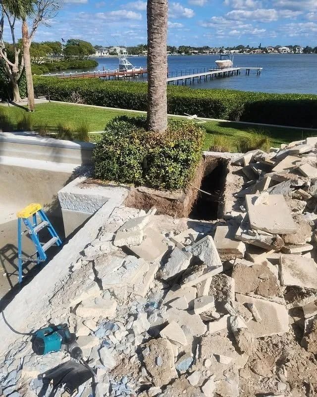 A construction site with debris and a ladder near a pool, with a palm tree and a waterfront view in the background.