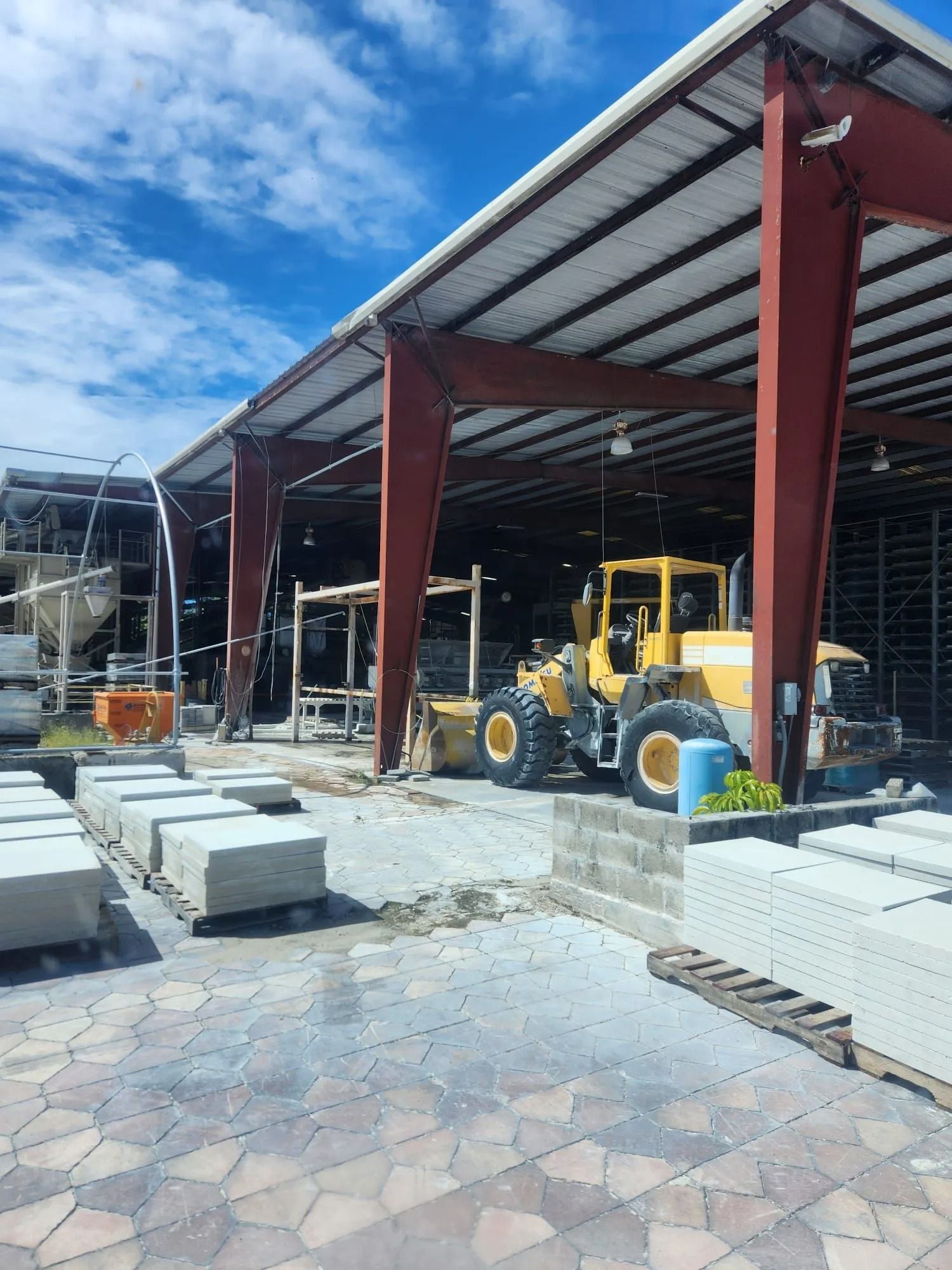 A yellow front-end loader parked under a large metal canopy at a concrete supply yard with stacks of paving stones.