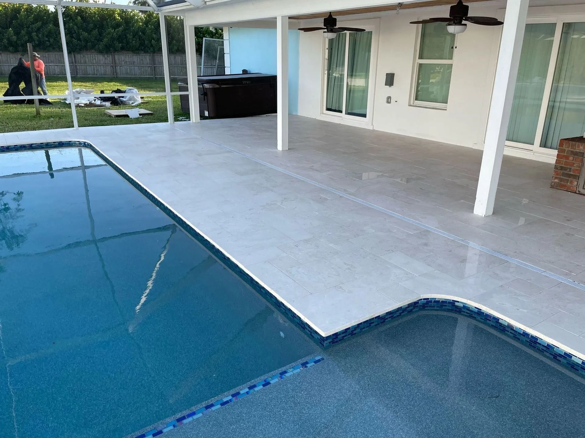 An outdoor pool deck with white pavers, blue tiled pool edges, and a covered patio area with ceiling fans.