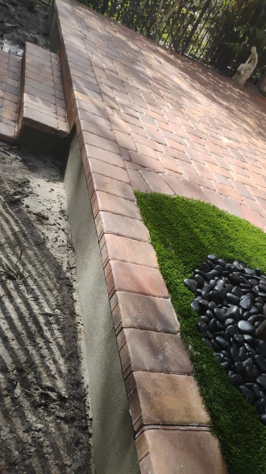 An elevated stone patio borders a patch of artificial grass and black river rocks, with a staircase leading up to the top.