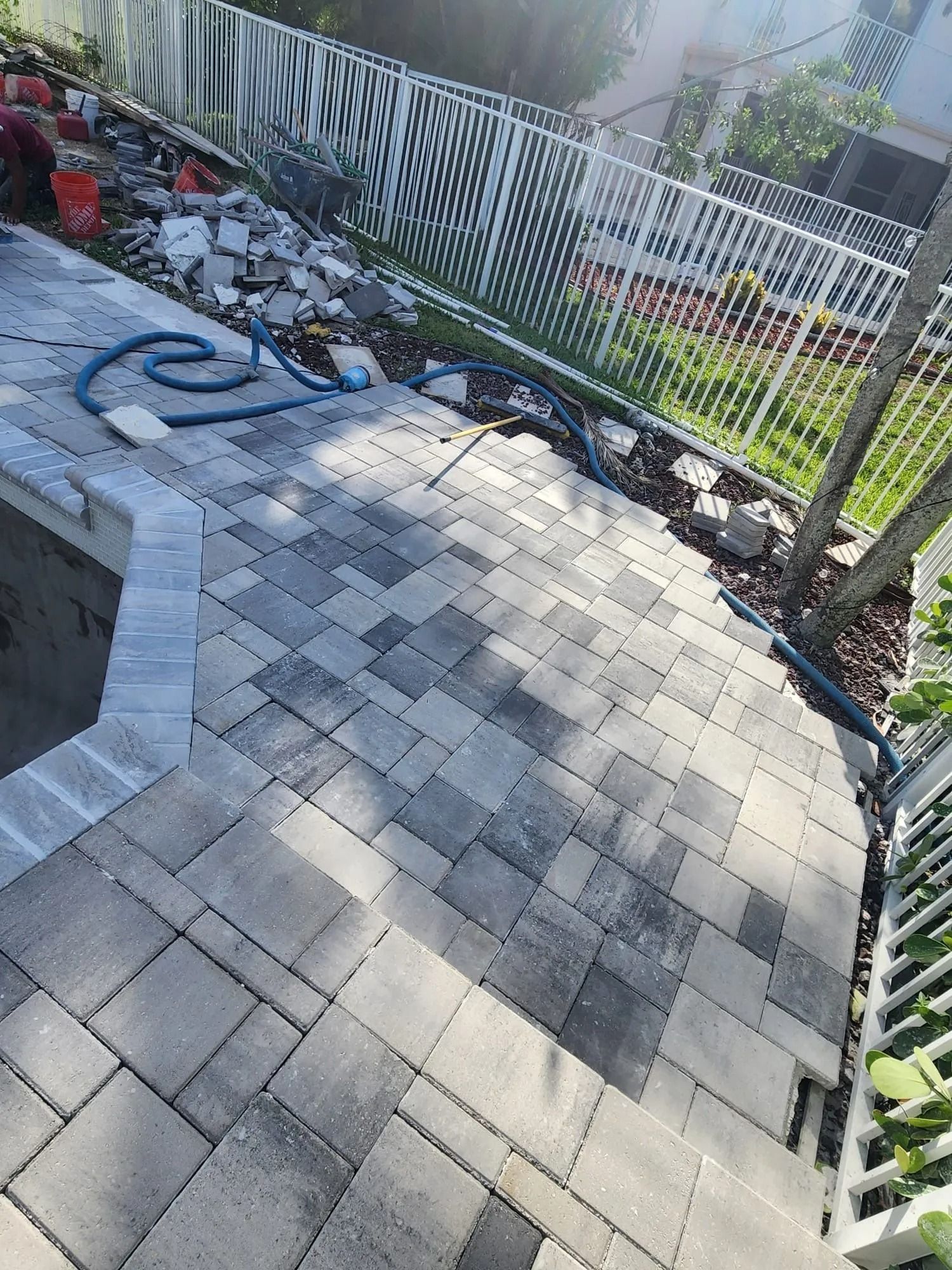 A poolside patio featuring gray, multi-sized pavers in a brick-style pattern, partially completed near a white fence.