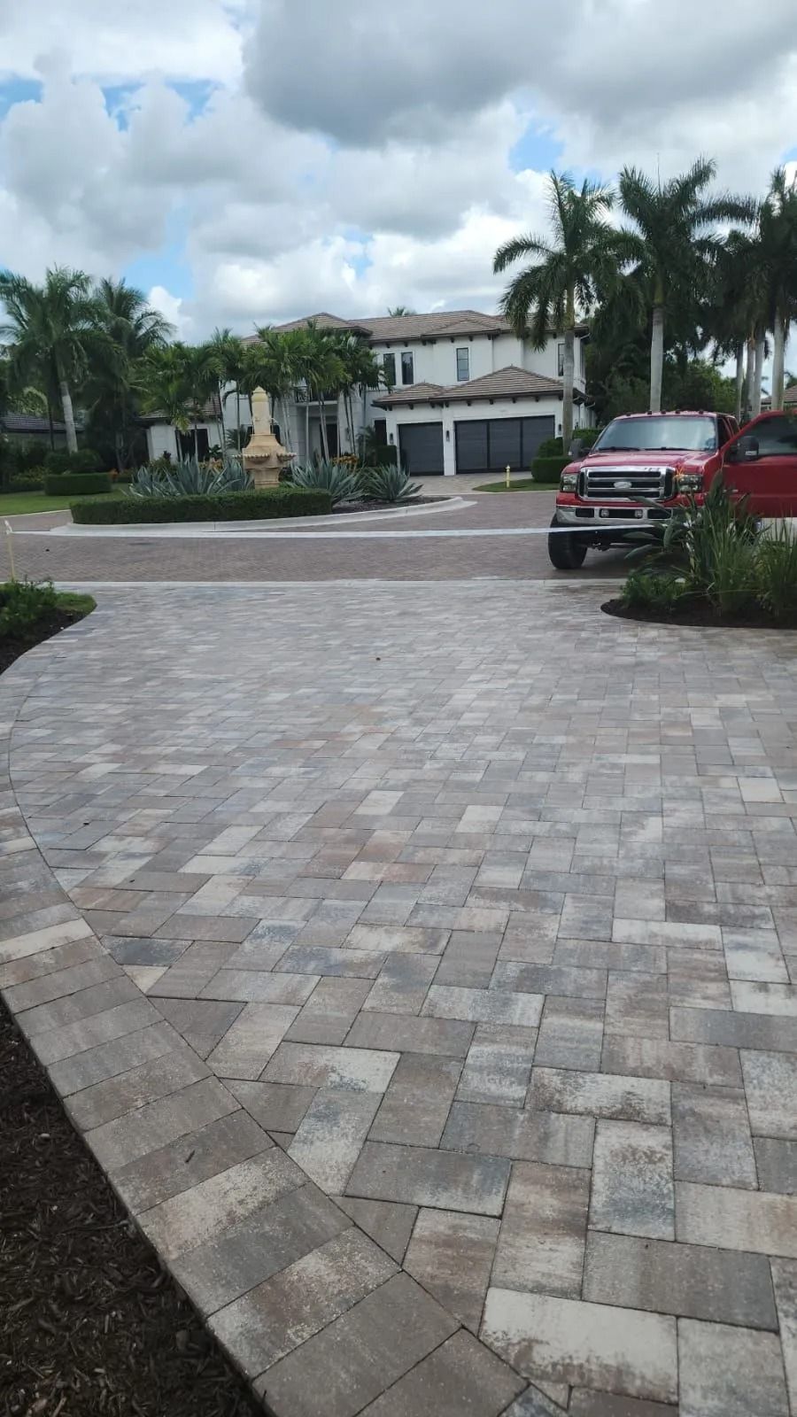 A paved driveway leads toward a two-story home with palm trees under a cloudy sky, with a red truck parked in the yard.