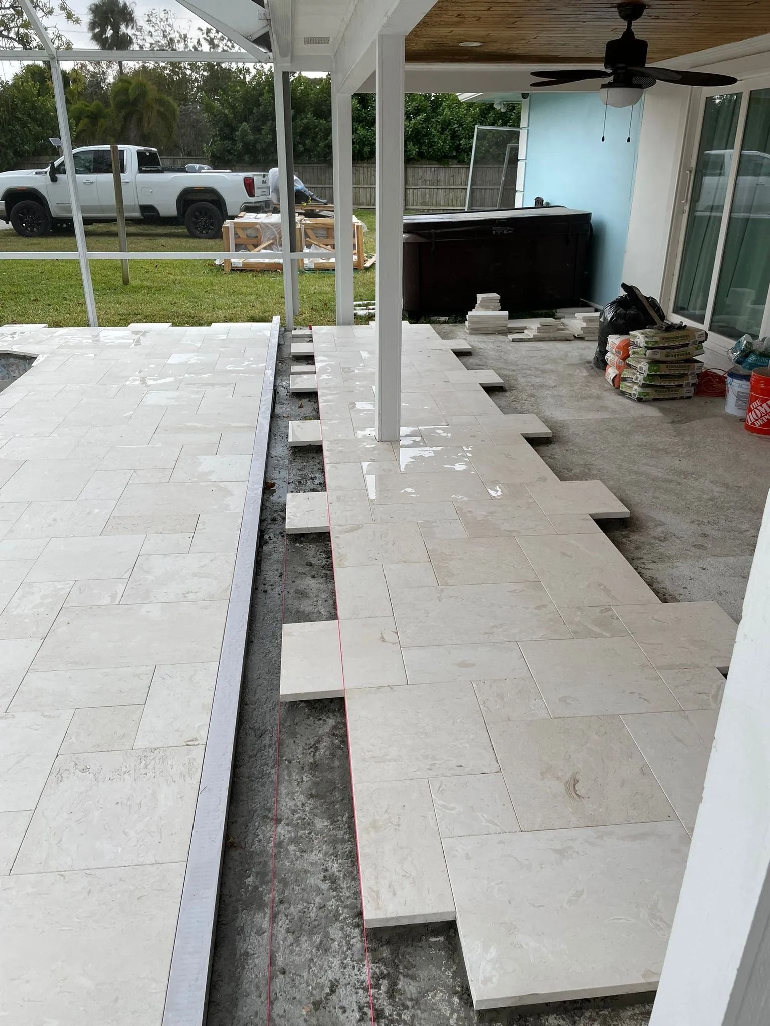A patio installation in progress with light-colored stone pavers being laid on a gravel base under a covered lanai.