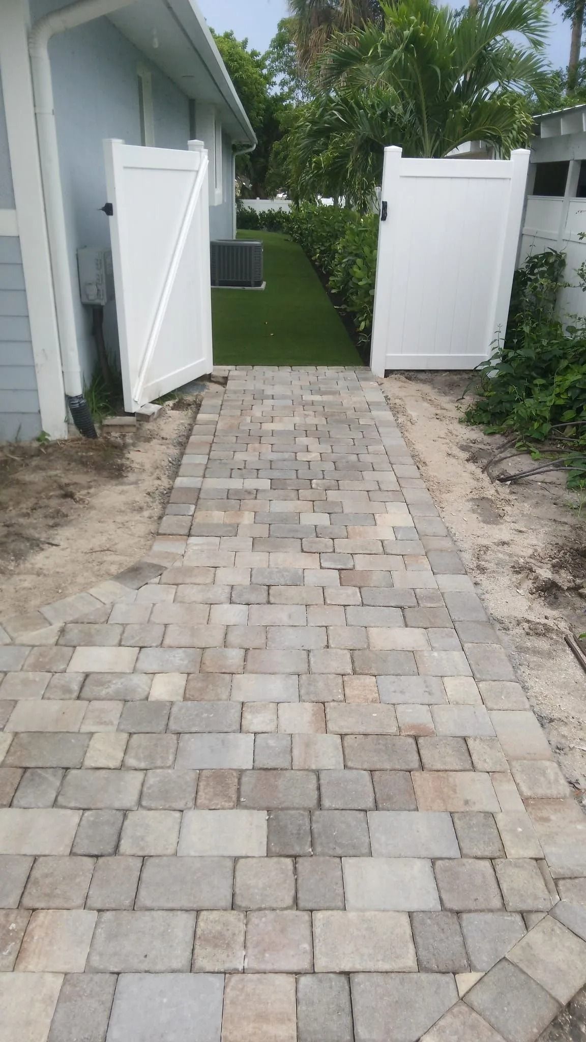 A paved stone walkway leading between two white vinyl fence gates to a green lawn area beside a house.