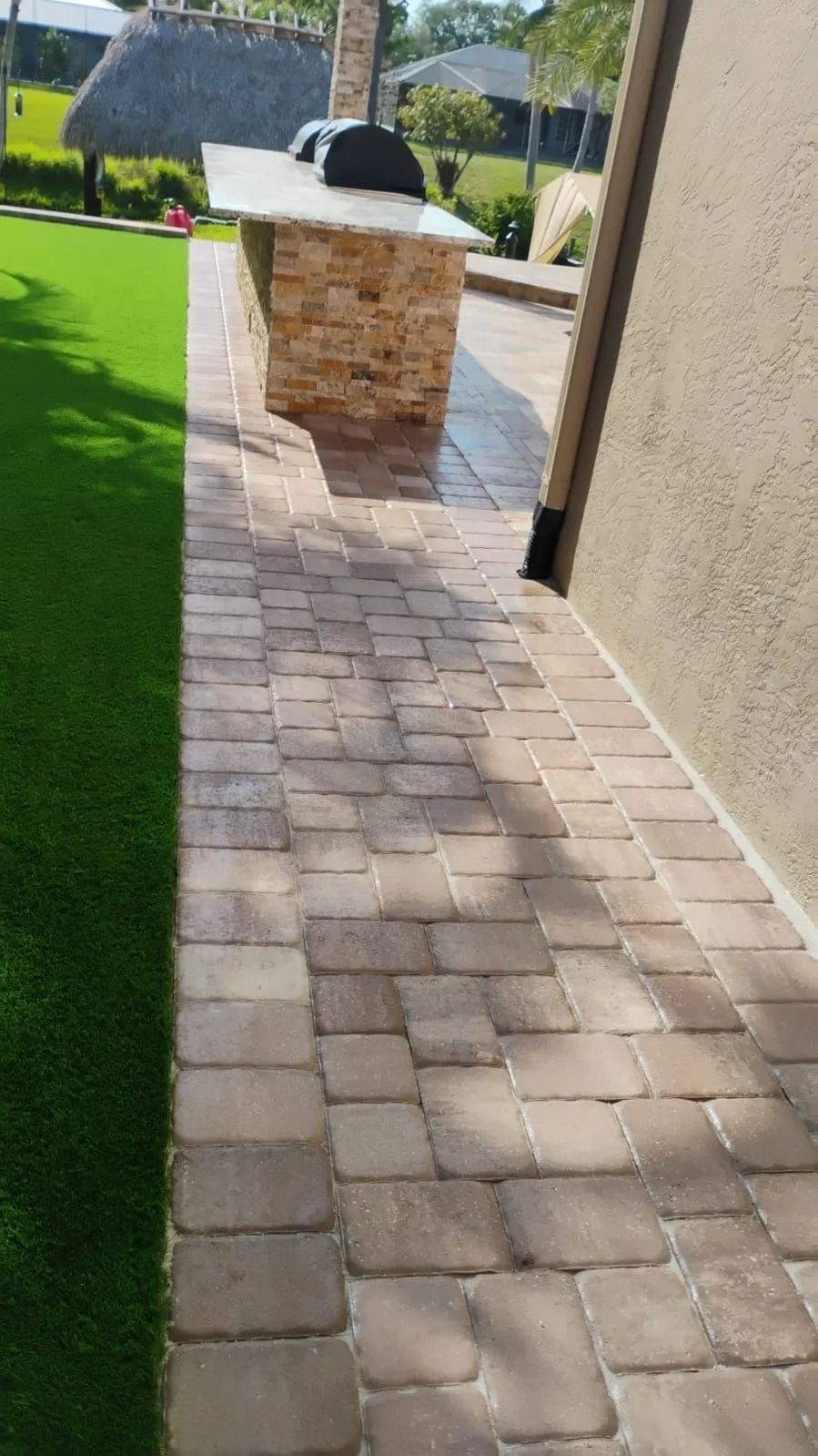 A stone patio pathway leads past a matching stone outdoor grill island beside a manicured green lawn and beige wall.