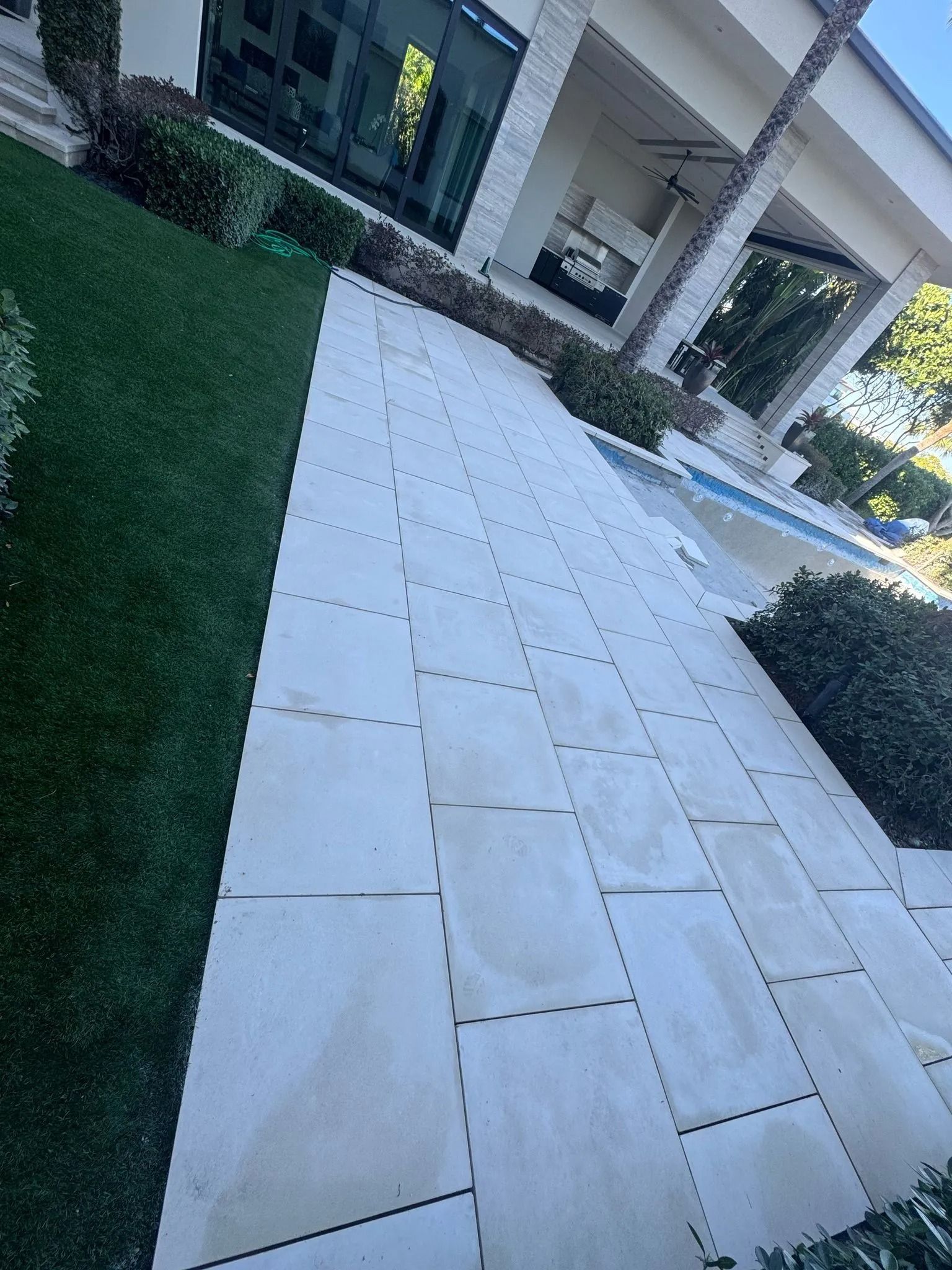 A light-colored stone patio leads toward a swimming pool, adjacent to a green lawn and modern building architecture.