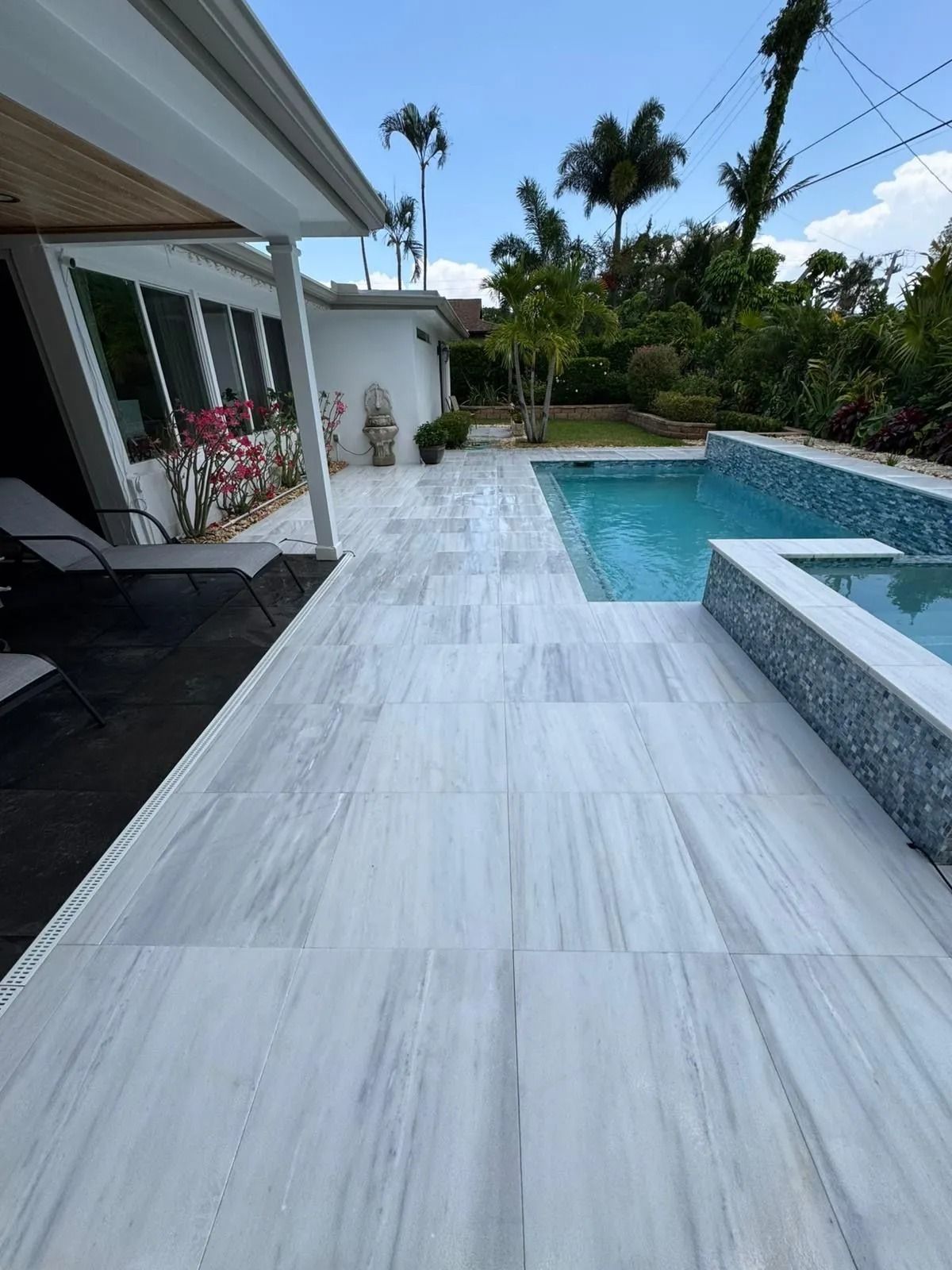 A bright outdoor patio with light gray stone tiles, a swimming pool, a hot tub, and a white house under a sunny blue sky.