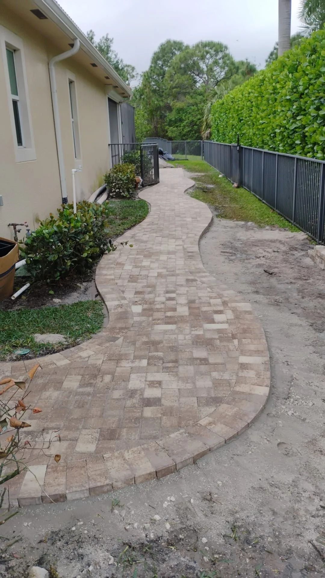 A winding brick paver walkway runs along the side of a house toward a green hedge and fence.