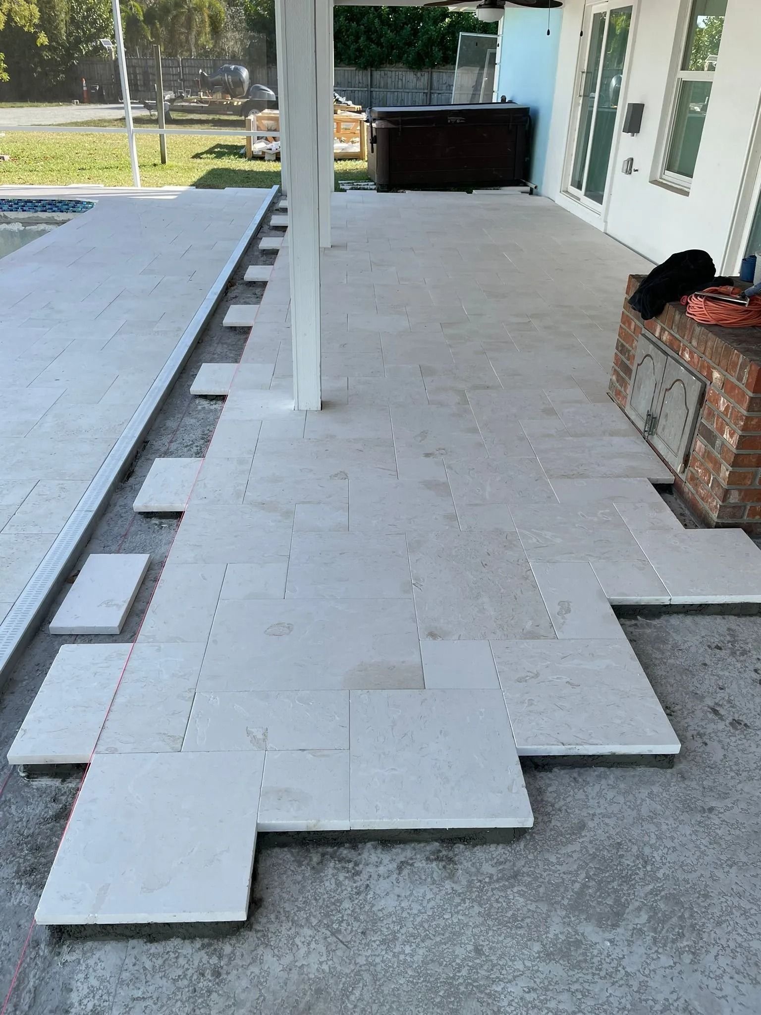 Light-colored stone pavers are being installed in a staggered pattern on a patio next to a swimming pool and brick wall.