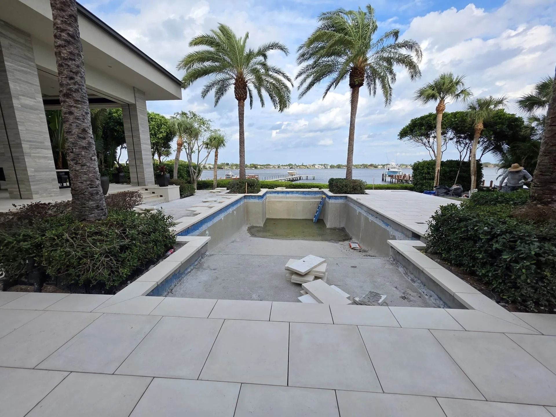 An empty, unfinished rectangular pool on a patio overlooking a calm waterfront with several palm trees under a cloudy sky.