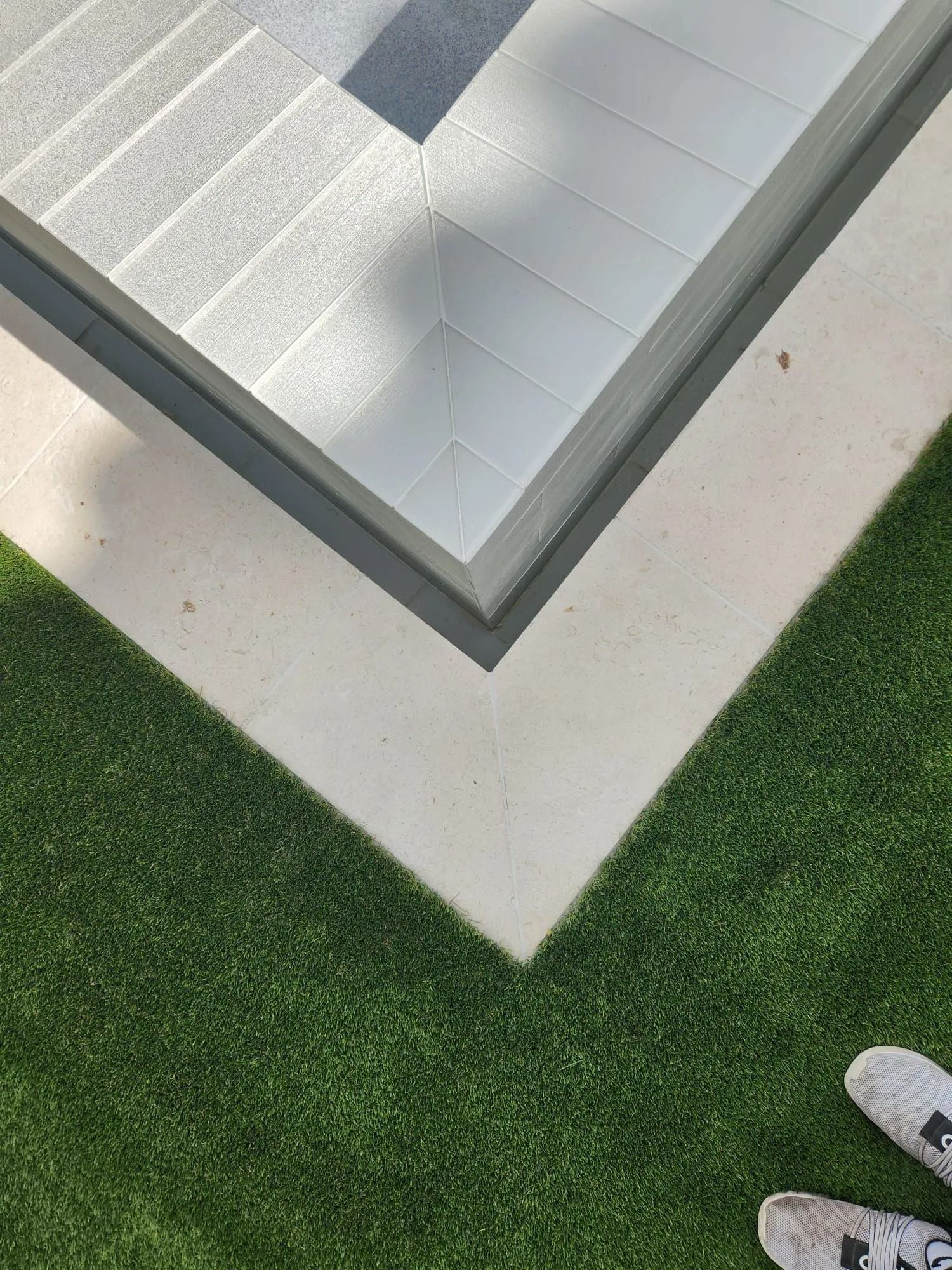 A top-down view of a sharp corner where gray decking, a beige stone border, and green artificial grass meet.