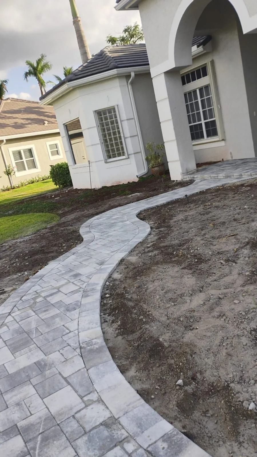 A grey paver walkway curves through a yard towards the arched entryway of a light-colored stucco home.